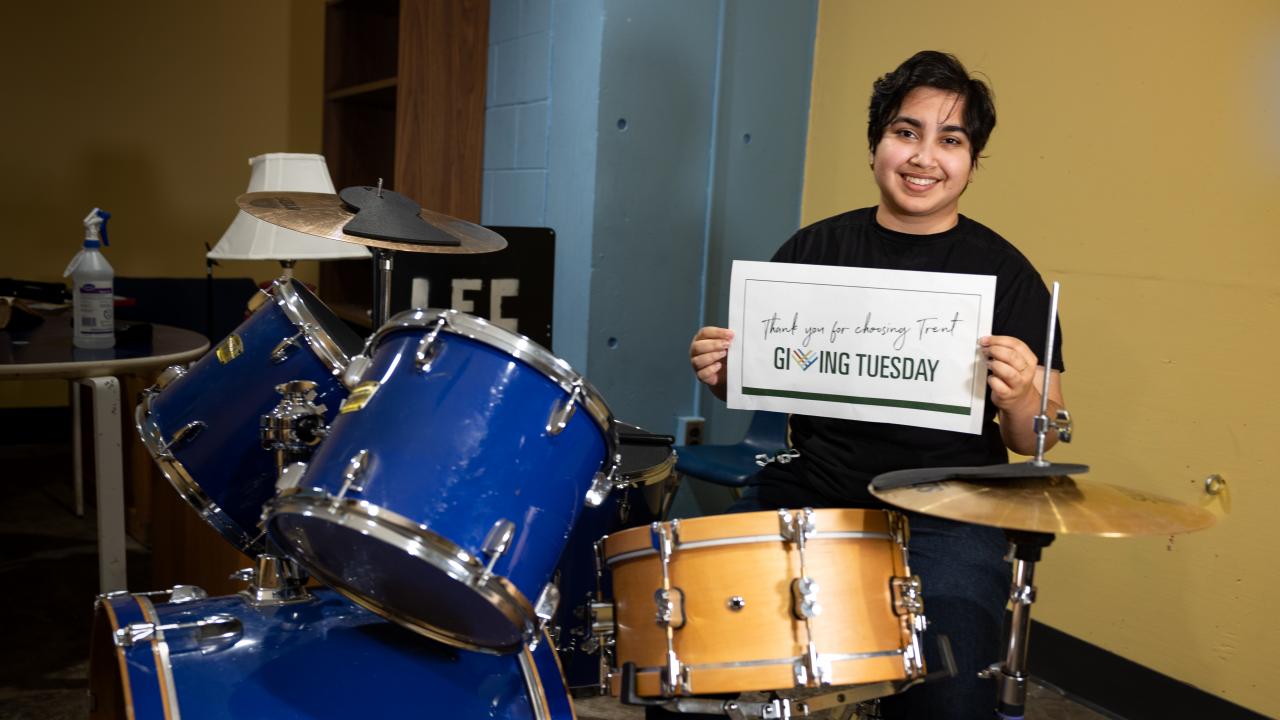Student smiling and holding giving Tuesday sign while sitting behind a drum set.
