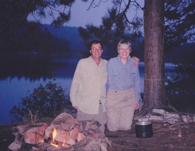 Susan and Duncan Gow camping - standing beside a campfire and in front of a lake. 